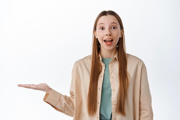 Giovane donna eccitata che tiene promo in mano aperta su copyspace sorridente prodotto di visualizzazione stupito in palmo allungato in piedi su sfondo bianco
