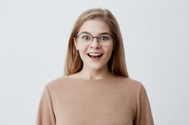 Giovane donna eccitata di aspetto europeo che indossa un maglione allentato marrone, con i capelli biondi, in occhiali, che sorride ampiamente per lo stupore, dimostrando i suoi denti bianchi perfetti. Gioventù e felicità