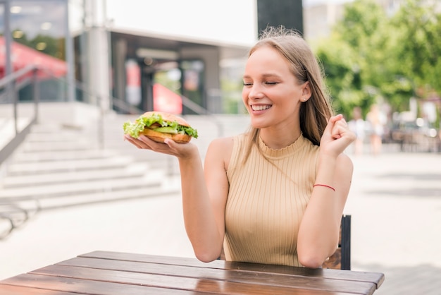 Giovane donna eccitata felice che tiene hamburger mentre è seduta all'aperto