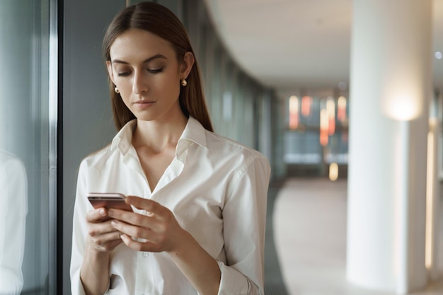 Giovane donna elegante in camicia bianca che controlla i messaggi che rispondono al business partner durante la pausa della riunione in piedi nel corridoio dell'ufficio utilizzando lo smartphone che manda un sms al collega che prova il discorso