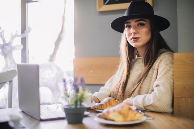 Giovane donna elegante in cappello con laptop e croissant al tavolo in caffetteria