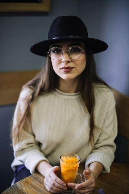 Giovane donna elegante in cappello ed occhiali con bicchiere di bevanda