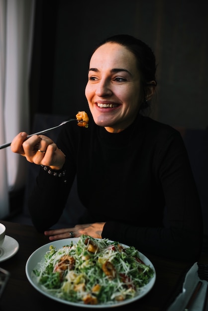Giovane donna elegante sorridente che mangia insalata alla tavola