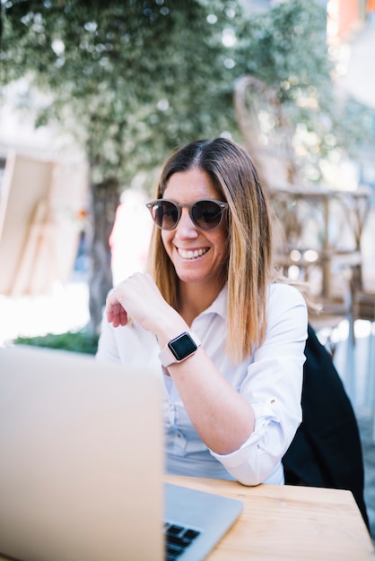 Giovane donna elegante sorridente che utilizza computer portatile alla tavola in caffè della via