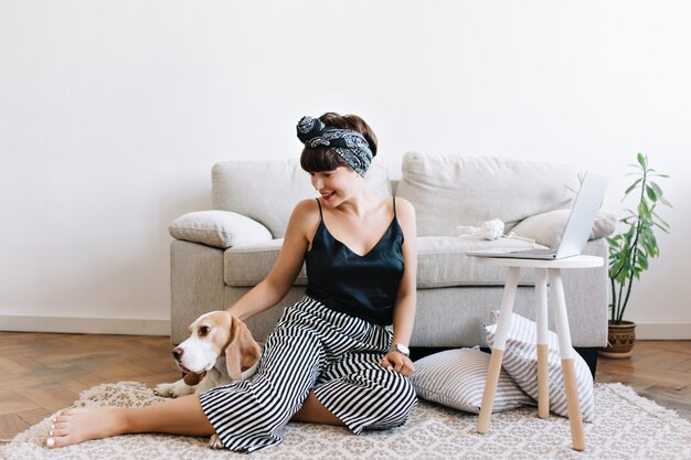 Giovane donna emozionante in pantaloni a strisce che si siedono sul pavimento accanto al sofà e al computer portatile che giocano con l'animale domestico