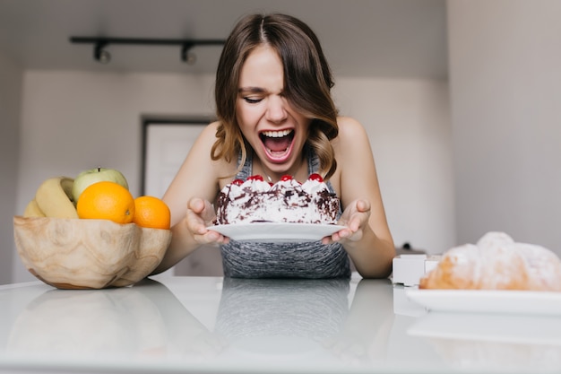 Giovane donna entusiasta che mangia torta cremosa. Modello femminile spensierato che gode di frutta e torta.