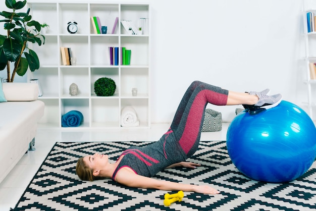 Giovane donna esile che si esercita con la palla blu dei pilates su tappeto a casa