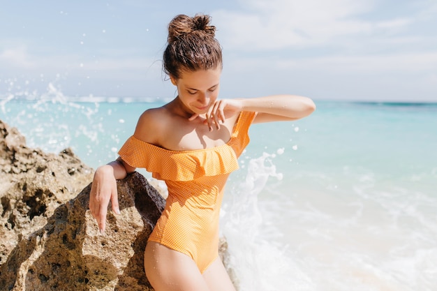 Giovane donna esile in un bellissimo costume da bagno giallo guardando verso il basso mentre posa in spiaggia. Magnifica ragazza caucasica che prende il sole in riva all'oceano.