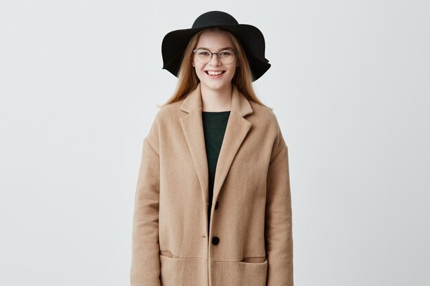 Giovane donna europea allegra con una bella serie di funzioni, vestita in cappotto sopra un maglione verde, indossa i suoi capelli biondi sciolti, con gli occhiali, guardando con un sorriso affascinante