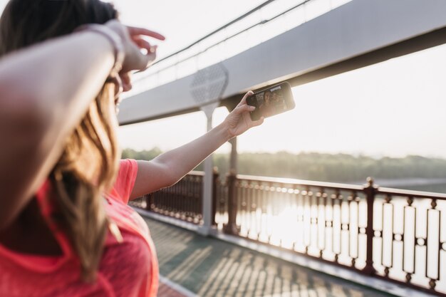 Giovane donna europea che fa selfie dopo l'allenamento. Ragazza allegra che cattura maschera di se stessa nella mattina soleggiata.