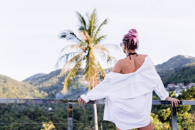 Giovane donna europea in bikini nero camicia lunga bianca con trecce rosa sulla testa su palme tropicali