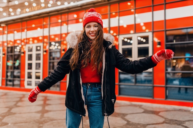 Giovane donna felice abbastanza sorridente in guanti rossi e cappello lavorato a maglia che indossa cappotto invernale a piedi in strada cittadina, vestiti caldi