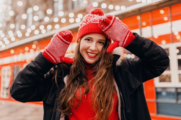 Giovane donna felice abbastanza sorridente in guanti rossi e cappello lavorato a maglia che indossa cappotto invernale che cammina nella strada di Natale della città, tendenza di moda stile vestiti caldi