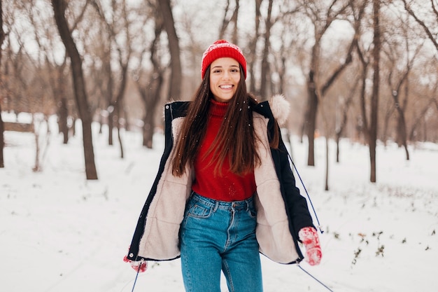 Giovane donna felice abbastanza sorridente in guanti rossi e cappello lavorato a maglia che indossa cappotto invernale, passeggiate nel parco nella neve, vestiti caldi