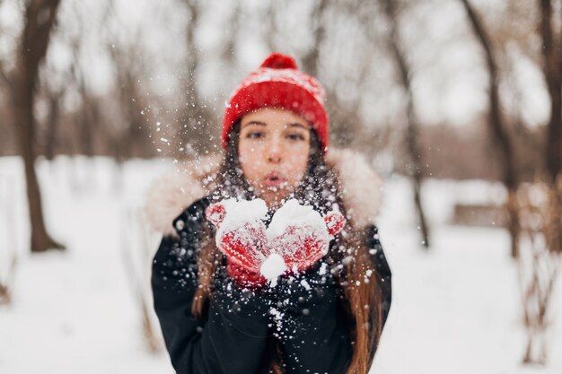 Giovane donna felice abbastanza sorridente in guanti rossi e cappello lavorato a maglia che indossa cappotto invernale, passeggiate nel parco, soffiando neve