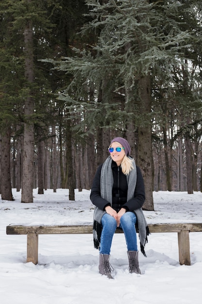 Giovane donna felice che si siede sul banco di legno che porta i vestiti caldi in inverno