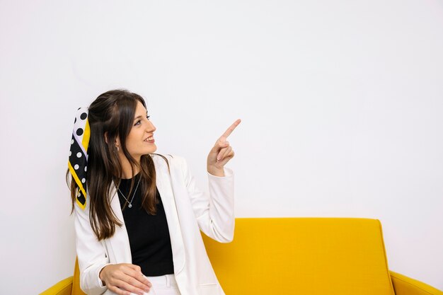 Giovane donna felice che si siede sul sofà giallo che indica verso l&#39;alto