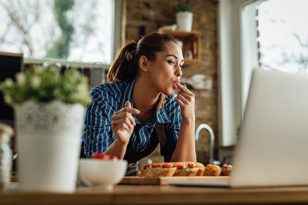 Giovane donna felice che usa il computer portatile mentre prepara la bruschetta in cucina.