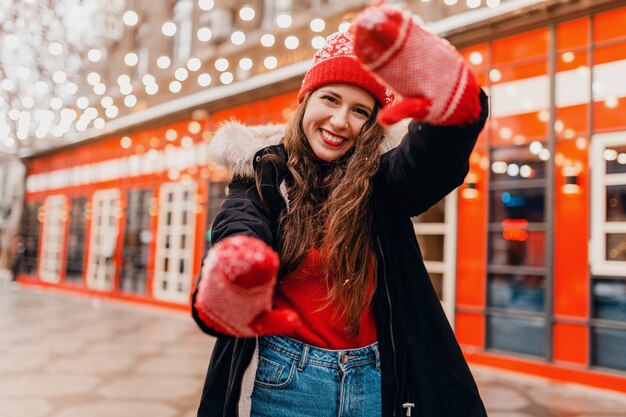 Giovane donna felice eccitata abbastanza sorridente in guanti rossi e cappello lavorato a maglia che indossa cappotto invernale che cammina nella strada di Natale della città, tendenza di moda stile vestiti caldi