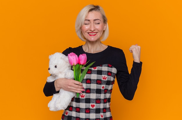 Giovane donna felice ed emozionata in vestito bello che tiene il mazzo di tulipani e orsacchiotto come regali che stringe il pugno che celebra la giornata internazionale della donna in piedi sopra il muro arancione