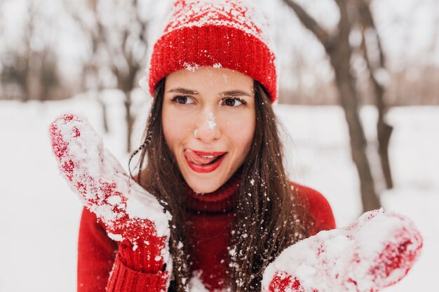 Giovane donna felice sorridente abbastanza candida con espressione faccia buffa in guanti rossi e cappello che indossa maglione lavorato a maglia a piedi giocando nel parco nella neve, vestiti caldi, divertendosi