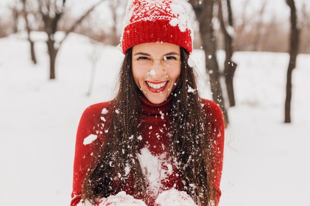 Giovane donna felice sorridente abbastanza candida in guanti rossi e cappello che indossa un maglione lavorato a maglia a piedi giocando nel parco nella neve, vestiti caldi, divertendosi