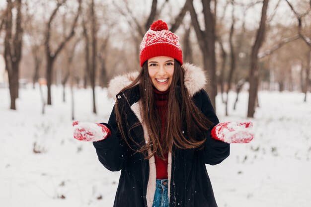 Giovane donna felice sorridente abbastanza candida in guanti rossi e cappello lavorato a maglia che indossa cappotto nero che cammina giocando nel parco nella neve, vestiti caldi, divertendosi