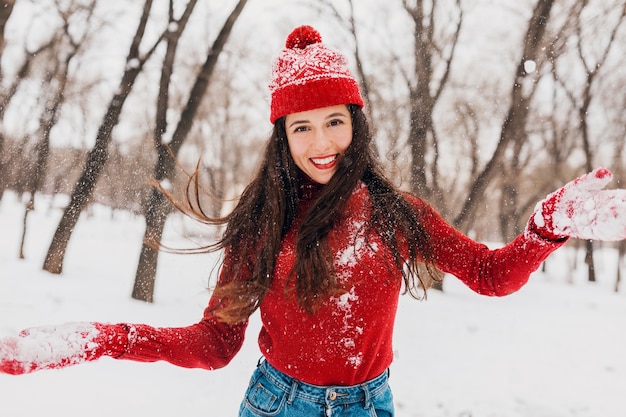 Giovane donna felice sorridente candida piuttosto eccitata in guanti rossi e cappello che indossa maglione lavorato a maglia a piedi giocando nel parco nella neve, vestiti caldi, divertendosi