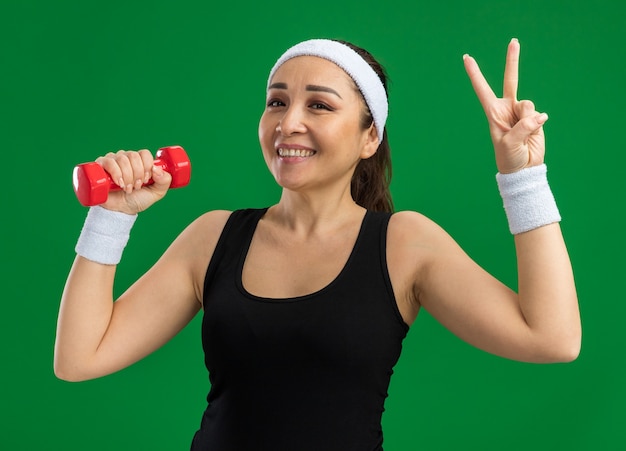 Giovane donna fitness con fascia con manubri che fa esercizi sorridendo mostrando il segno v in piedi sul muro verde green