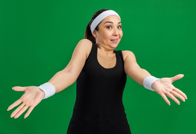 Giovane donna fitness con fascia e bracciali confusi con le braccia in segno di dispiacere e indignazione in piedi sul muro verde