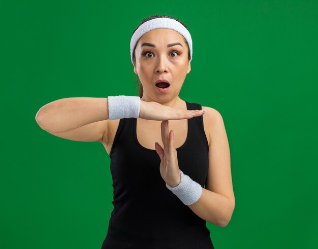 Giovane donna fitness con fascia e bracciali sorpresa a fare un gesto di pausa con le mani in piedi sul muro verde green