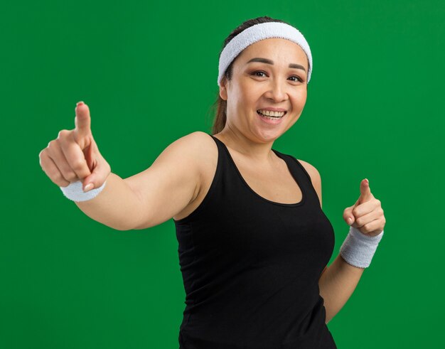 Giovane donna fitness con fascia e bracciali sorridenti fiduciosi che puntano con le dita indice in piedi sul muro verde