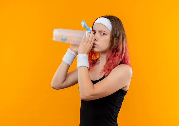 Giovane donna fitness in abbigliamento sportivo acqua potabile con viso serio in piedi sopra la parete arancione