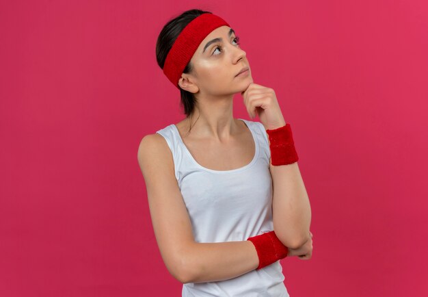 Giovane donna fitness in abbigliamento sportivo con archetto guardando da parte il pensiero perplesso in piedi sopra il muro rosa