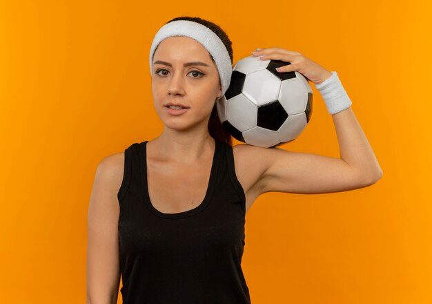 Giovane donna fitness in abbigliamento sportivo con fascia tenendo il pallone da calcio guardando fiducioso in piedi sopra la parete arancione