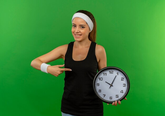 Giovane donna fitness in abbigliamento sportivo con fascia tenendo l'orologio da parete che punta con il dito ad esso sorridente fiducioso in piedi oltre la parete verde