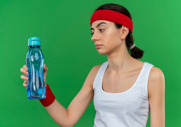 Giovane donna fitness in abbigliamento sportivo con fascia tenendo una bottiglia di acqua guardandolo scontento con l'espressione scettica in piedi sopra la parete verde