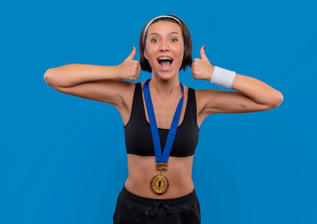 Giovane donna fitness in abbigliamento sportivo con medaglia d'oro al collo che mostra i pollici in su rallegrandosi del suo successo felice ed eccitato in piedi sopra la parete blu
