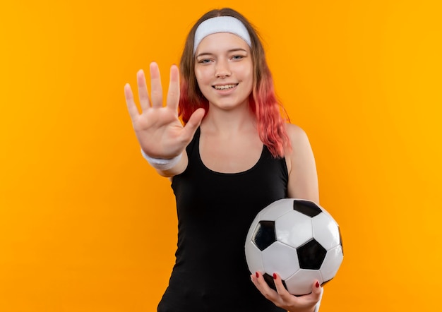 Giovane donna fitness in abbigliamento sportivo tenendo il pallone da calcio facendo il segnale di stop con la mano, sorridente in piedi sopra il muro arancione