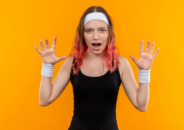 Giovane donna fitness in abiti sportivi che sembra sorpreso alzando le mani nella resa in piedi sopra la parete arancione