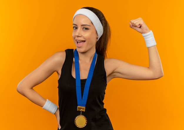 Giovane donna fitness in abiti sportivi con fascia e medaglia d'oro intorno al collo alzando il pugno sorridendo felice e positivo in piedi sopra la parete arancione