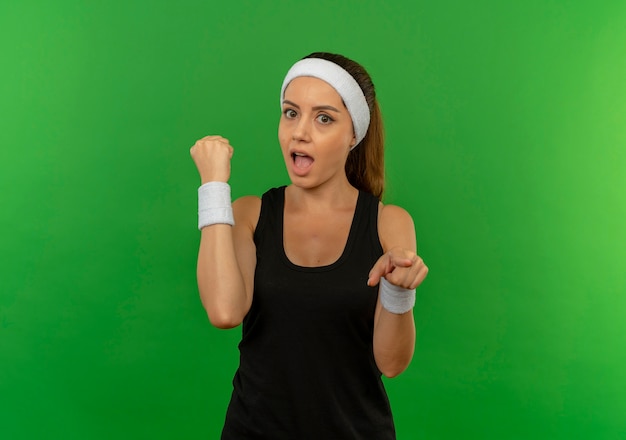 Giovane donna fitness in abiti sportivi con fascia pugno di serraggio puntato con il dito alla telecamera guardando sorpreso in piedi sopra la parete verde