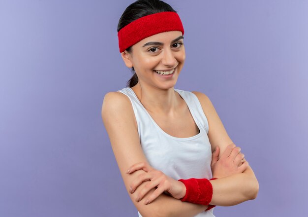 Giovane donna fitness in abiti sportivi con fascia sorridendo allegramente con le braccia incrociate sul petto in piedi sopra la parete viola