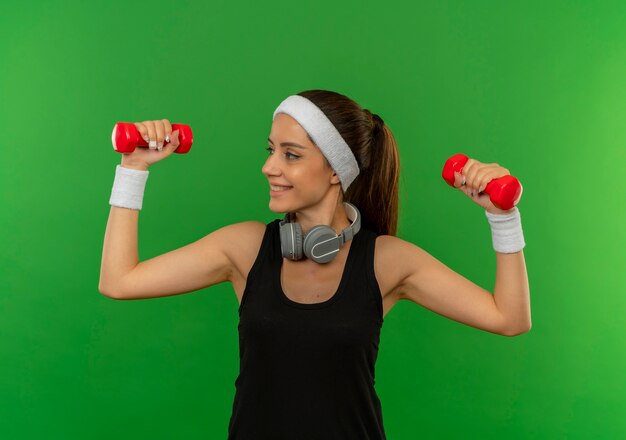 Giovane donna fitness in abiti sportivi con fascia tenendo due manubri facendo esercizi guardando fiducioso con il sorriso sul viso in piedi sopra la parete verde