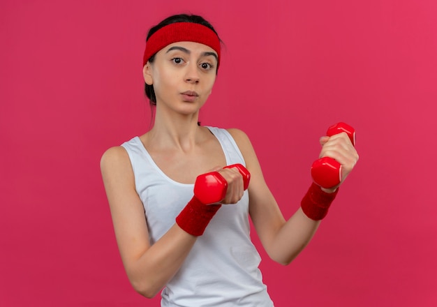 Giovane donna fitness in abiti sportivi con fascia tenendo due manubri in mani alzate guardando fiducioso facendo esercizi in piedi sopra il muro rosa