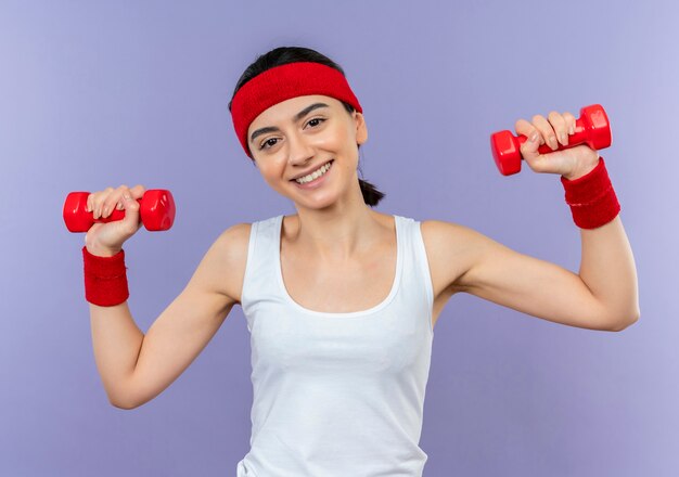 Giovane donna fitness in abiti sportivi con fascia tenendo due manubri in mani alzate sorridendo allegramente facendo esercizi in piedi sopra la parete viola