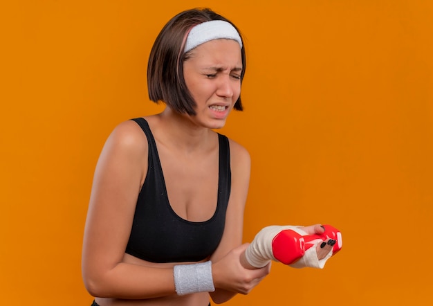 Giovane donna fitness in abiti sportivi con fascia tenendo il manubrio toccando il polso che sembra malessere sensazione di dolore in piedi sopra la parete arancione