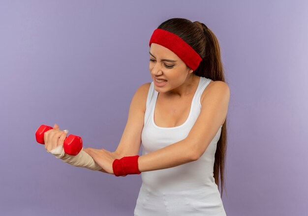 Giovane donna fitness in abiti sportivi con fascia tenendo il manubrio toccando il suo braccio bendato guardando malessere in piedi sopra il muro grigio