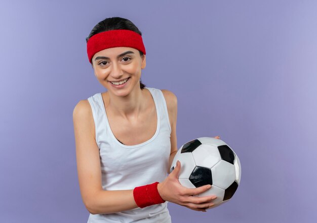 Giovane donna fitness in abiti sportivi con fascia tenendo il pallone da calcio con il sorriso sul viso in piedi sopra la parete viola