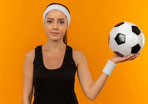 Giovane donna fitness in abiti sportivi con fascia tenendo il pallone da calcio con la faccia seria in piedi sopra la parete arancione